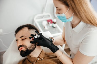 Stylish bearded man visiting aesthetic clinic, getting lips filler, closeup. Man having beauty injection at male spa salon. Anti-aging treatment for men concept