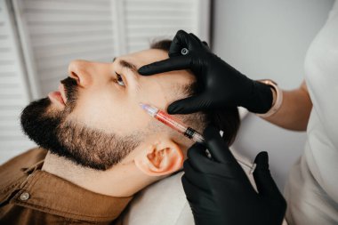 Beauty treatments for a stylish young man with a beard. Beautician makes beauty injections in a man's face