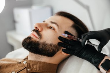 Stylish bearded man visiting aesthetic clinic, getting lips filler, closeup. Man having beauty injection at male spa salon. Anti-aging treatment for men concept