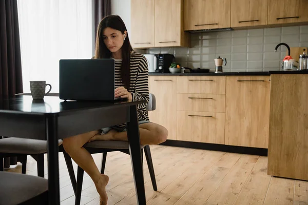 Freelancer woman sits by the table in the home kitchen office, working on laptop. Playful child distracts from work, kid making noise and asking attention from busy mom