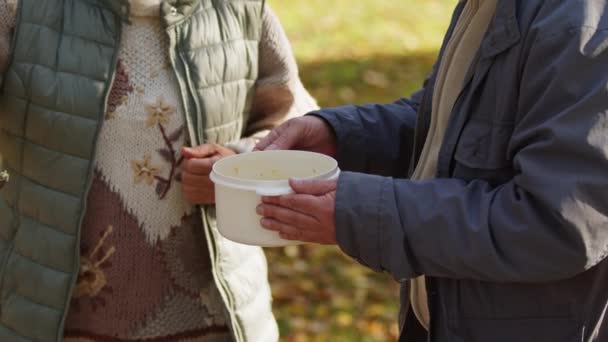 Социальное обеспечение. Благотворительное мероприятие. Женщина наливает горячий суп в пластиковый контейнер в руки старика — стоковое видео