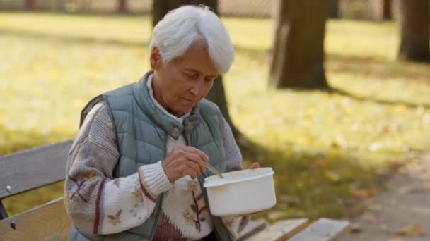 Γριά γυναίκα με γκρίζα μαλλιά τρώει το γεύμα στο πάρκο από πλαστικό δοχείο το φθινόπωρο — Αρχείο Βίντεο
