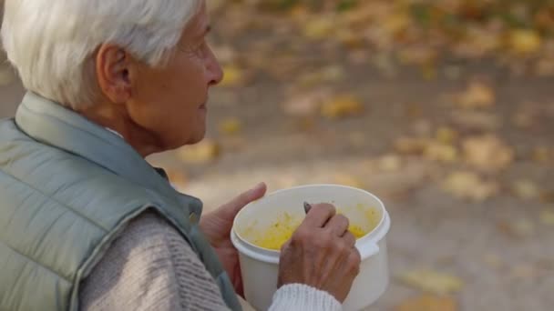 Traurige einsame Frau mit grauen Haaren isst im Herbst im Park Essen aus Plastikbehältern — Stockvideo