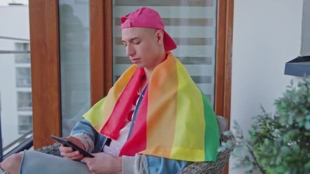 Homosexuální muž pokrytý duhovou vlajkou pomocí tabletu, zatímco sedí na terase — Stock video