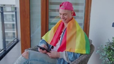 Terasta otururken tablet kullanan, gökkuşağı bayraklarıyla kaplı gülümseyen homoseksüel bir adam.