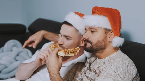 Όμορφο γκέι ζευγάρι με καπέλα Σάντα που τρώνε πίτσα και βλέπουν ταινία παραμονή Χριστουγέννων. — Φωτογραφία Αρχείου