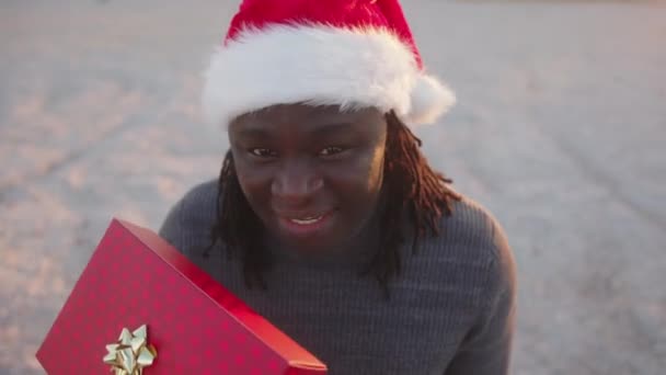 Szczęśliwy czarny człowiek w kapeluszu Mikołaja trzyma czerwony zapakowany prezent świąteczny. Strzał pod wysokim kątem — Wideo stockowe