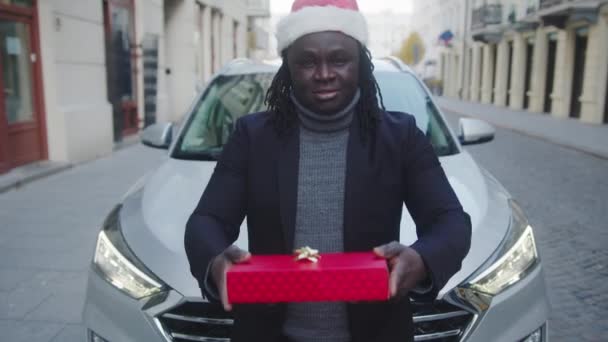 Jeune homme noir avec chapeau de Père Noël donnant rouge enveloppé cadeau de Noël devant sa voiture — Video