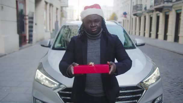 Молодой черный мужчина в шляпе Санты дарит красный рождественский подарок перед своей машиной — стоковое видео