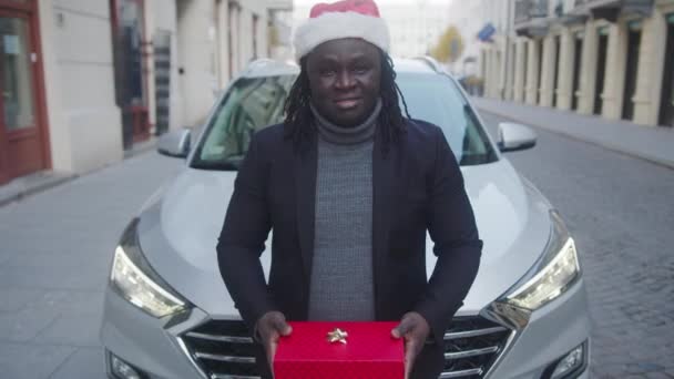 Jeune homme noir avec chapeau de Père Noël donnant rouge enveloppé cadeau de Noël devant sa voiture — Video