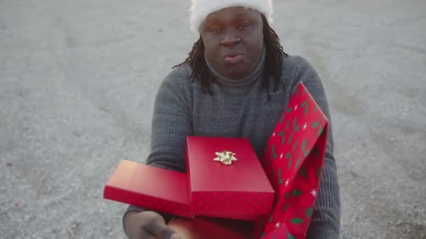 Czarny człowiek w kapeluszu Mikołaja trzyma kilka czerwonych opakowanych prezentów świątecznych — Wideo stockowe
