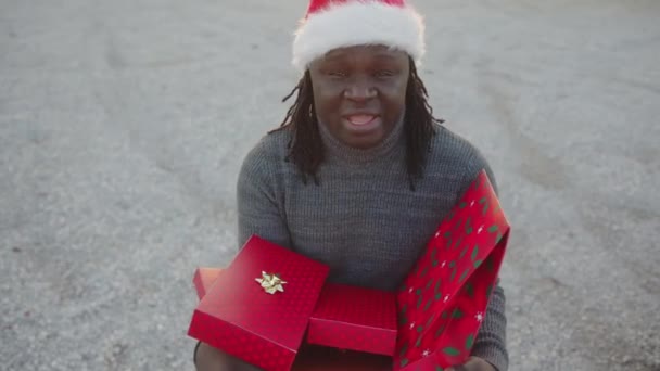 Svart man med tomte hatt håller gäng röda inslagna julklappar — Stockvideo