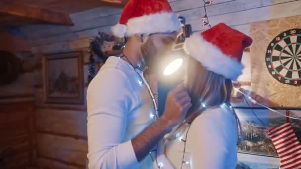 Feliz pareja joven bailando con luces navideñas y sombreros de santa. Amor y afecto durante la víspera de Navidad — Vídeo de stock