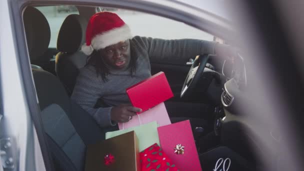 Bonito homem negro sentado no carro com chapéus de Papai Noel cercado por presentes de Natal — Vídeo de Stock