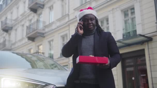 Африканский американец в шляпе Санты и с подарком разговаривает по телефону перед машиной — стоковое видео