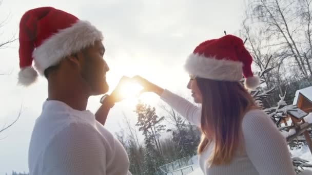 Świąteczny duch. Młoda para z kapeluszami Mikołaja całuje się trzymając kształt serca oprawiając słońce na zaśnieżonej górze — Wideo stockowe
