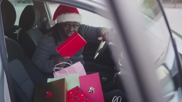 Bonito homem negro sentado no carro com chapéus de Papai Noel cercado por presentes de Natal — Vídeo de Stock