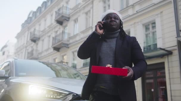 Noel şapkası ve kırmızı hediye kutusu olan siyahi bir adam şehir caddesinde telefon görüşmesi yapıyor. — Stok video