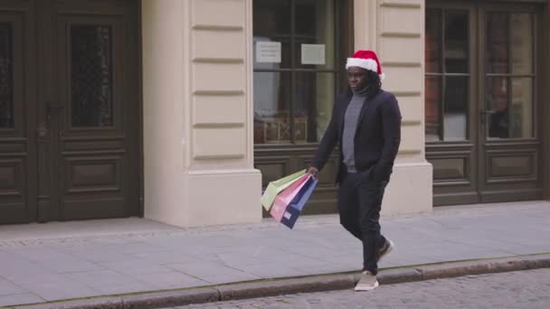 Czarny człowiek w świątecznym kapeluszu spacerujący po ulicy z prezentami świątecznymi — Wideo stockowe