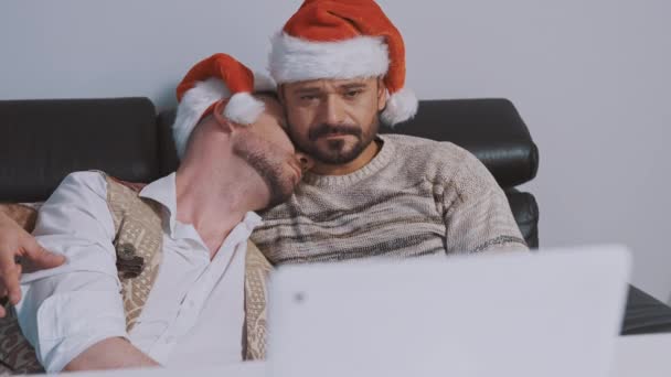 Молодая гей-пара с санта-хатами обнимается и смотрит фильмы накануне Рождества — стоковое видео