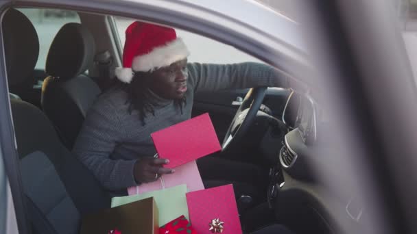 Bonito homem negro sentado no carro com chapéus de Papai Noel cercado por presentes de Natal — Vídeo de Stock