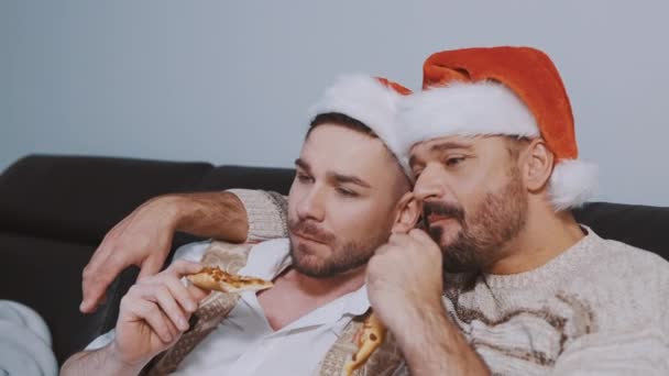 Krásný gayové párek s vánoční klobouky jíst pizzu a sledování filmů na Štědrý večer — Stock video