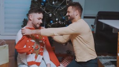 Yeni evli eşcinsel çiftin ilk Noel 'i. Adam Noel ağacının önünde ortağına Noel süveteri veriyor.