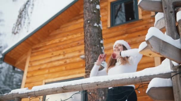 Jonge blanke vrouw met kerst hoed genieten van sneeuw vallen op het balkon van een houten huis — Stockvideo