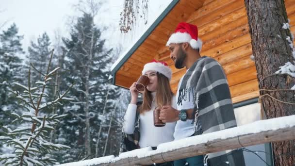 Kerstochtend. Gelukkig jong stel drinken warm drankje in de voorkant van het houten huis op de berg bedekt met sneeuw — Stockvideo