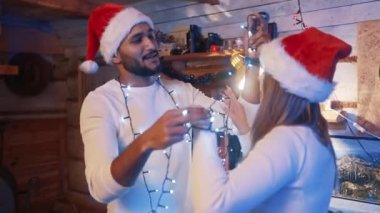 Noel arifesinde Noel Baba şapkaları ve yılbaşı ışıklarıyla dans eden mutlu, çok ırklı çift.