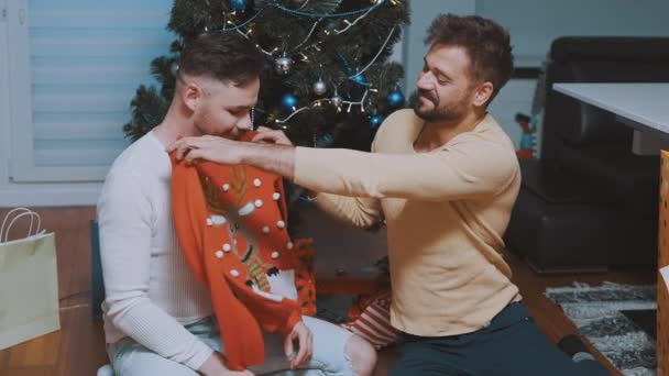 Первое Рождество молодоженов-геев. Мужчина дарит рождественский свитер своему партнеру перед елкой — стоковое видео