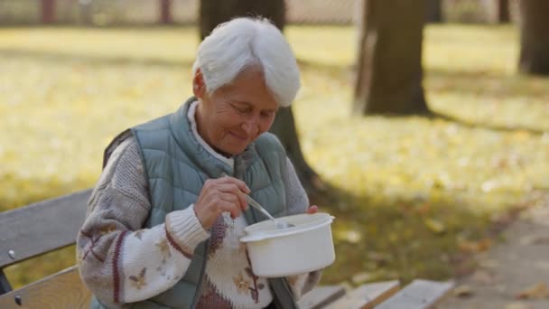 Пожилая женщина ест благотворительную еду, сидя на скамейке в парке — стоковое видео