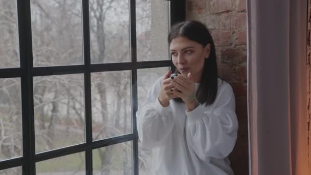 Giovane donna caucasica che beve caffè mattutino guardando attraverso la finestra in inverno — Video Stock