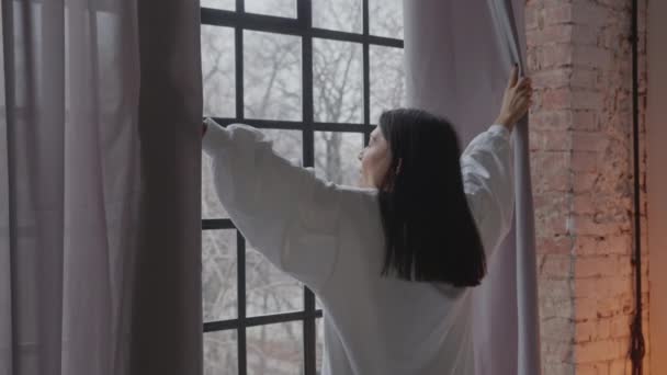 Ung kvinna håller gardiner och tittar genom fönstret. Glad självsäker dam njuter av vacker utsikt och drömmer hemma — Stockvideo