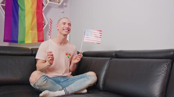 Ευτυχισμένος νεαρός ομοφυλόφιλος βλέποντας τηλεόραση κρατώντας αμερικανικές σημαίες - μακρινό σουτ — Αρχείο Βίντεο