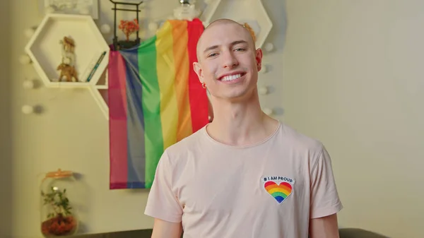 Libero uguale felice uomo gay caucasico sorridente alla telecamera, in posa nel suo appartamento, e in piedi di fronte alla bandiera dell'orgoglio arcobaleno. — Foto Stock