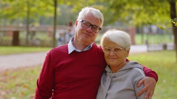 Пожилой муж-пенсионер обнимает любимую жену и улыбается в камеру. Средний крупный план на открытом воздухе. — стоковое видео