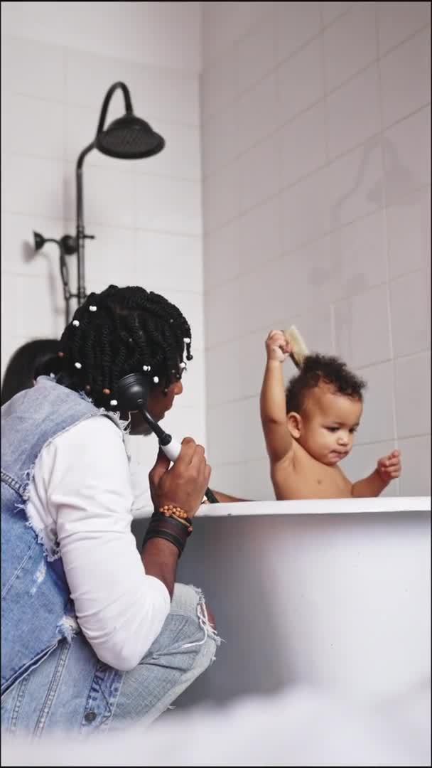 Ένα μωρό μικτής φυλής που διασκεδάζει σε μια μπανιέρα. Αφρο-Αμερικανός πατέρας και το παιδί του. Κάθετη εσωτερική λήψη. — Αρχείο Βίντεο