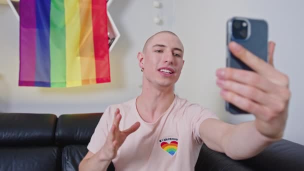 Гордий лисий гей-інфлюенсер знімає собі відео зі смартфоном, райдужний прапор гордості на задньому плані . — стокове відео