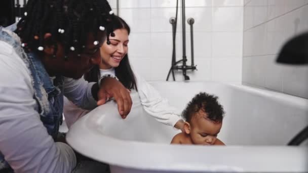 Interraciaal paar zitten in de badkamer met hun schattige peuter in de badkuip spelen met douchekop. — Stockvideo