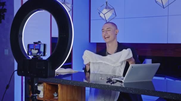 Caucasiano gay influenciador em seu 20s criação novo moda haul para seus seguidores online, mostrando branco malha t-shirt e falando sobre roupas — Vídeo de Stock
