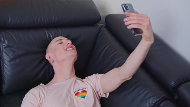 Quirky divertido caucásico queer influencer toma divertidos selfies con cámara frontal mientras se acuesta en un sofá negro. — Vídeo de stock