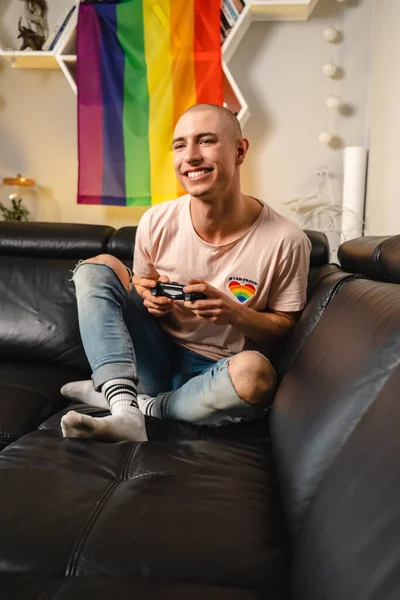 Amigável feliz queer gay ativista jogar vídeo games com seu gamepad enquanto sentado no um preto sofá. Tiro interior vertical. — Fotografia de Stock