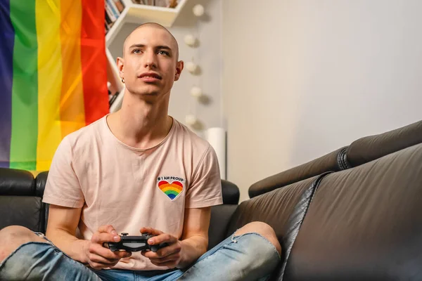 Careca queer ativista caucasiano usando gamepad e tentando ganhar um jogo. Bandeira do arco-íris no fundo. — Fotografia de Stock