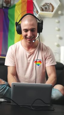 Siyah bir kanepede otururken dizüstü bilgisayarında bilgisayar oyunu oynayan kel, beyaz eşcinsel bir oyuncu. İçeriden dikey çekim. 