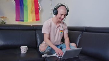 Arka planda gökkuşağı bayrağı olan bir kanepede otururken, kulaklıklı, kulaklık takan tuhaf, kel adam..