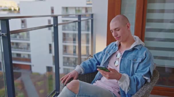 Молодий квір-підприємець використовує смартфон, сидячи на балконі, насолоджуючись гарною погодою і свіжим повітрям . — стокове відео