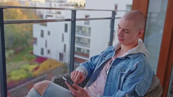 Självsäker flintskallig bög kille sitter på en fåtölj på sin balkong, ser båda sidor, koppla av, med hjälp av smartphone. — Stockvideo