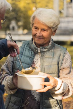 Avrupalı dost canlısı gri saçlı yaşlı kadın, ihtiyacı olan insanlarla parkta lezzetli sıcak çorbasını paylaşıyor..