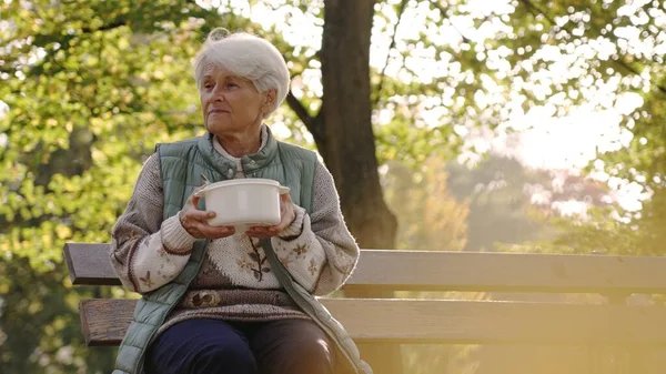 Caucasian elderly homeless woman sitting on a bench and holding a plastic bowl filled with hot soup given to her by volunteers.